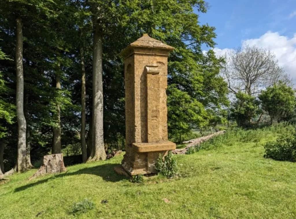 Cromwell's Seat, a tall Cotswold limestone monument with a stone seat niche on a grassy hillside beside woodland