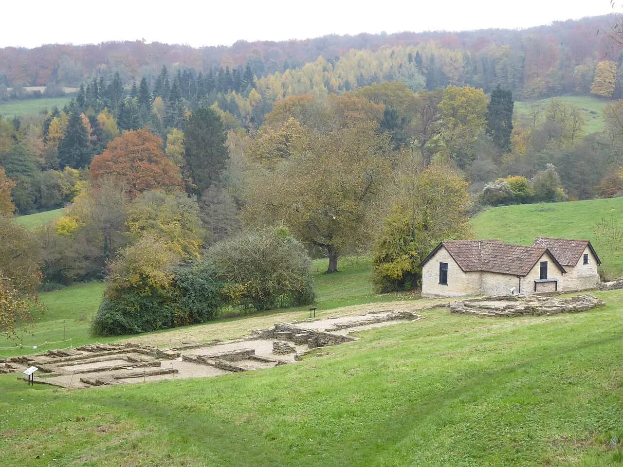 Stone foundations of Great Witcombe Roman Villa on a grassy slope, with a small protective building and autumn woodland beyond