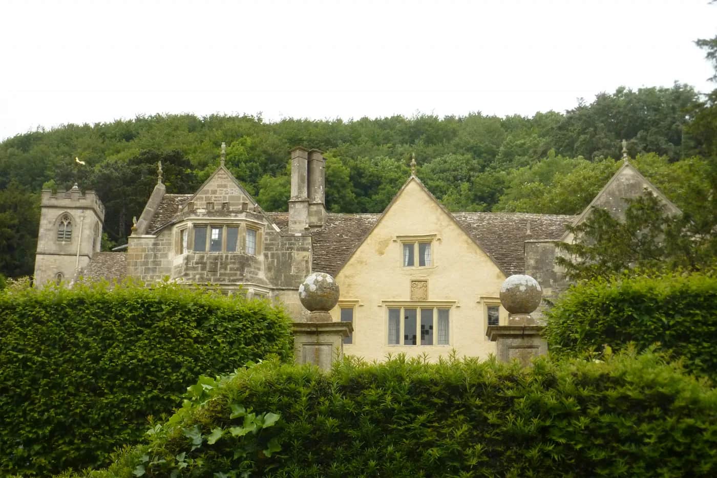 Front facade of Owlpen Manor, a multi-gabled Tudor Cotswold-stone house with mullioned windows, stone-slate roofs, and tall chimneys set against a wooded hillside