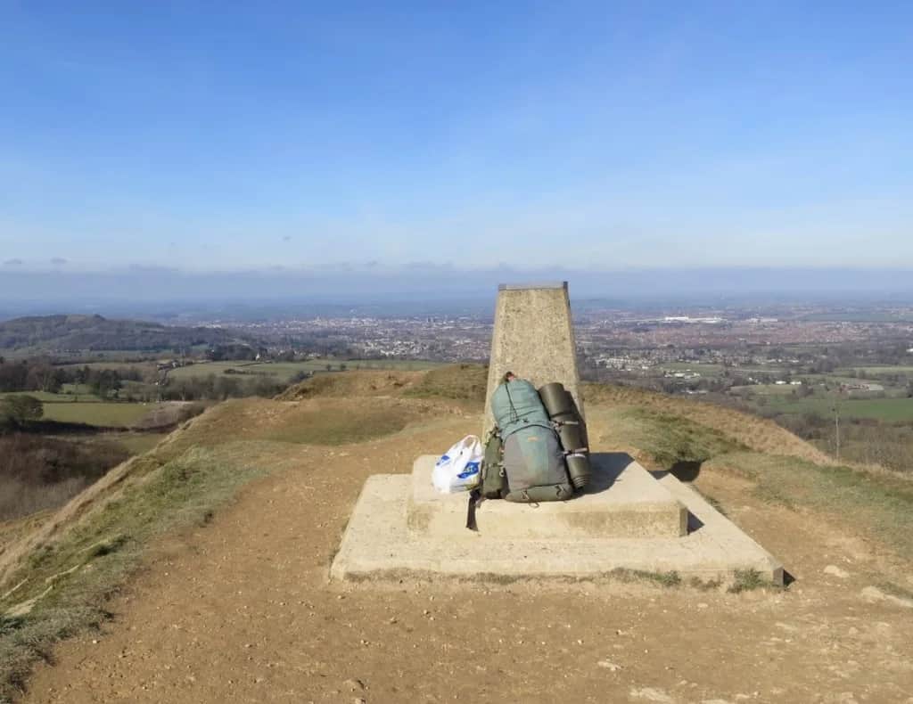 Trig point on Painswick Beacon with a hiker's pack, overlooking the Severn Vale toward Gloucester