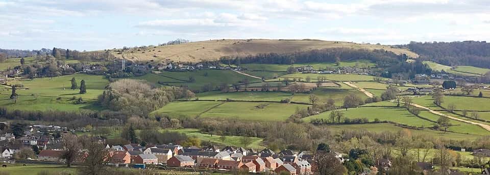 Panoramic view of the flat-topped grassy plateau of Selsley Common rising above green fields and housing in the Stroud valleys