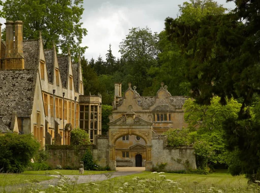 Stanway House and its ornate Cotswold stone gatehouse, seen across lawns and wildflowers beneath woodland