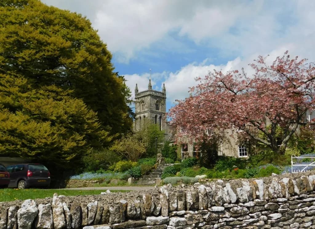 A Cotswold village scene with a stone church tower, cherry blossom, and a dry stone wall