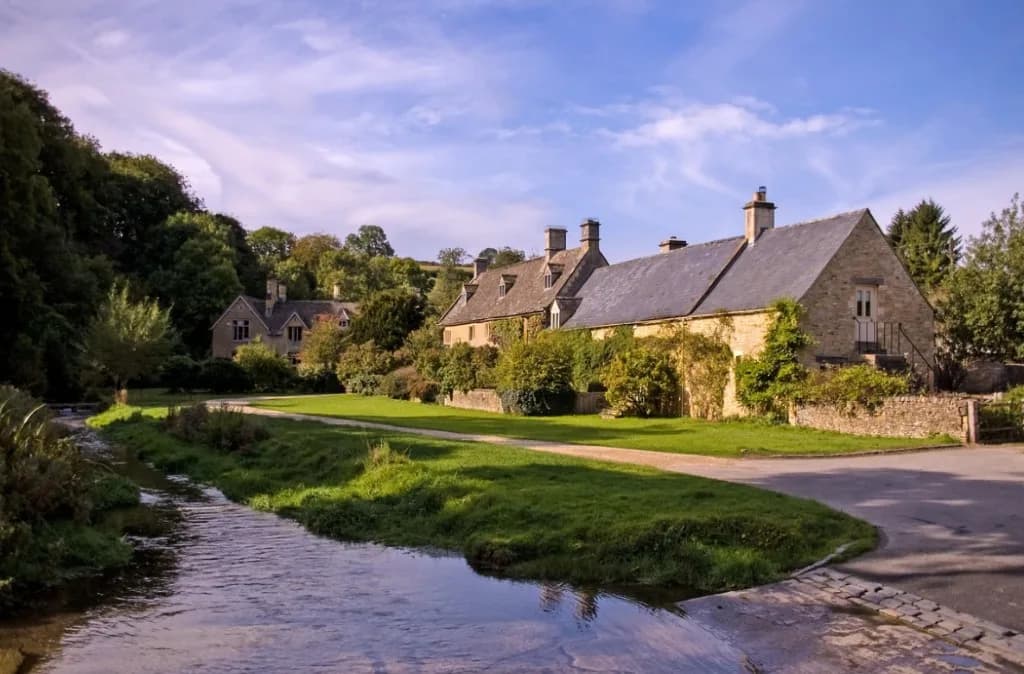 Honey-coloured Cotswold stone cottages beside a stream in a green village setting