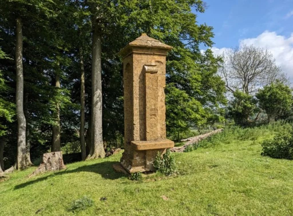 Cromwell's Seat, a tall Cotswold limestone monument with a stone seat niche on a grassy hillside beside woodland