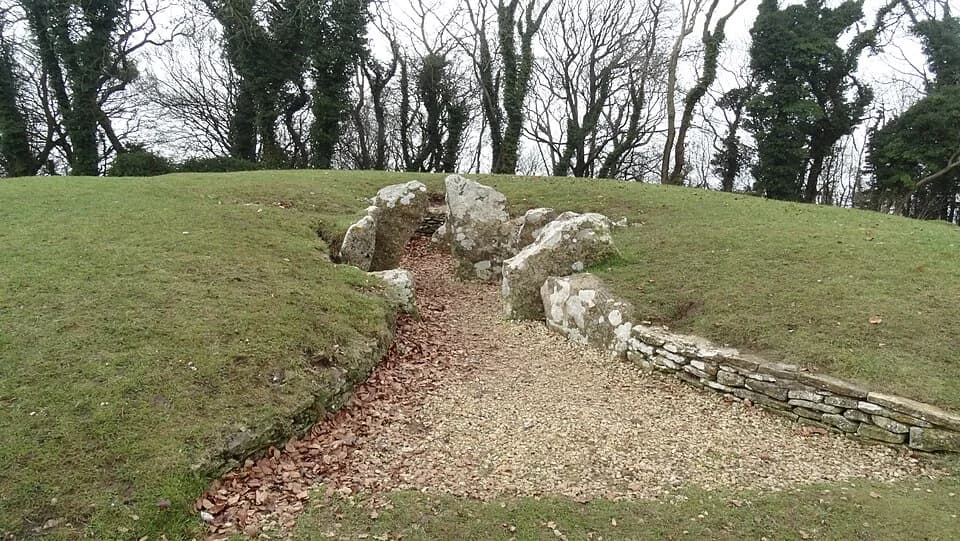 Stone entrance and chambers of Nympsfield Long Barrow set into a grassy mound with bare winter trees beyond