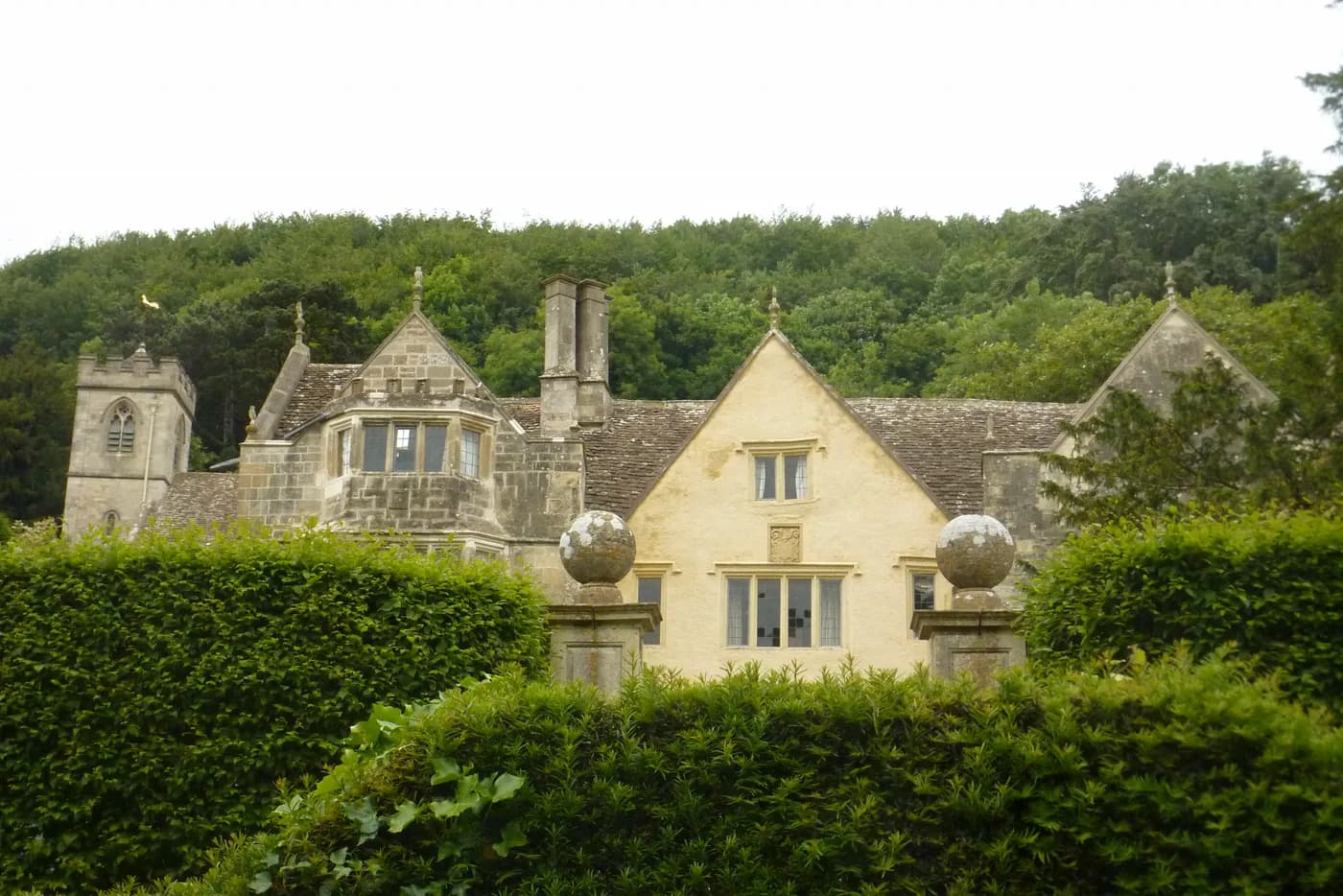 Front facade of Owlpen Manor, a multi-gabled Tudor Cotswold-stone house with mullioned windows, stone-slate roofs, and tall chimneys set against a wooded hillside