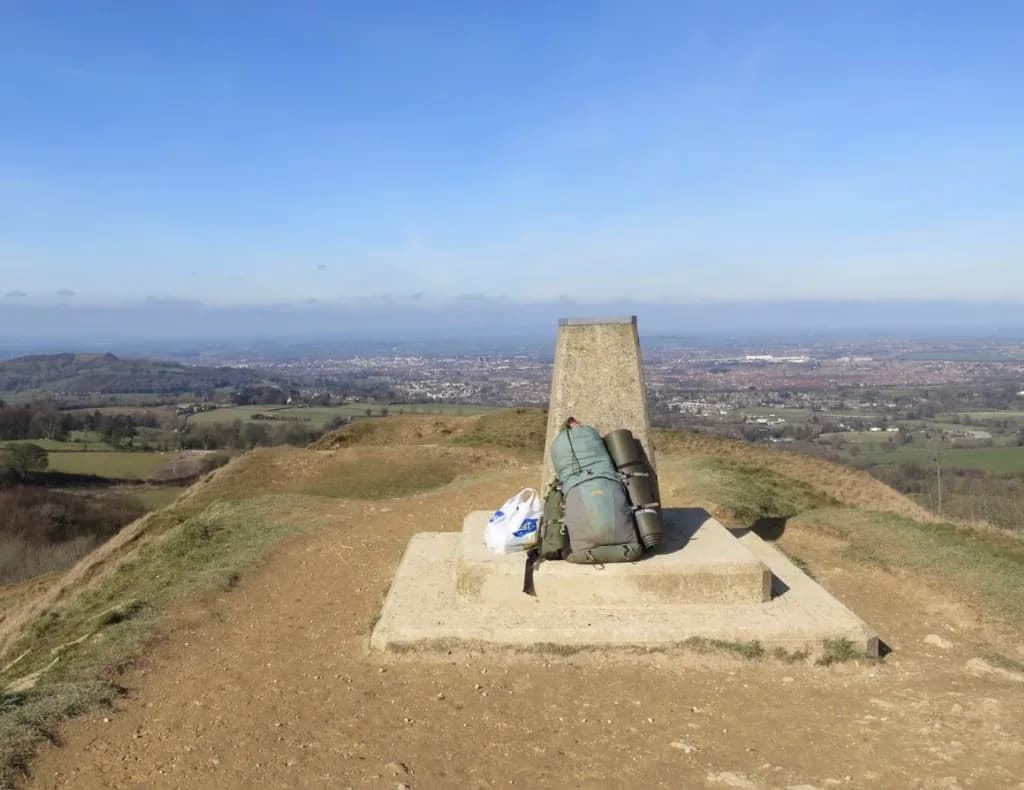 Trig point on Painswick Beacon with a hiker's pack, overlooking the Severn Vale toward Gloucester