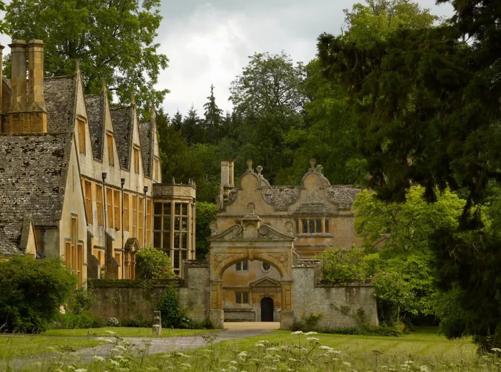Stanway House and its ornate Cotswold stone gatehouse, seen across lawns and wildflowers beneath woodland